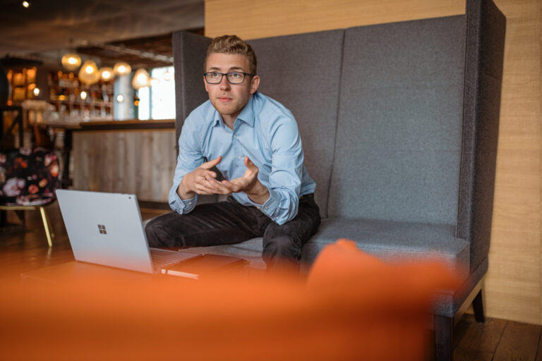 Mann im hellblauen Hemd, der auf einem grauen Sofa sitzt und gestikuliert, während er auf einen Laptop schaut. Er spricht über datengetriebenes Webdesign, Website-Check, Website-Analyse und Website-Optimierung in einer gemütlichen, modernen Umgebung. Der Fokus liegt auf der Analyse des Nutzerverhaltens und der Entwicklung von Optimierungsvorschlägen zur Verbesserung der Website-Performance